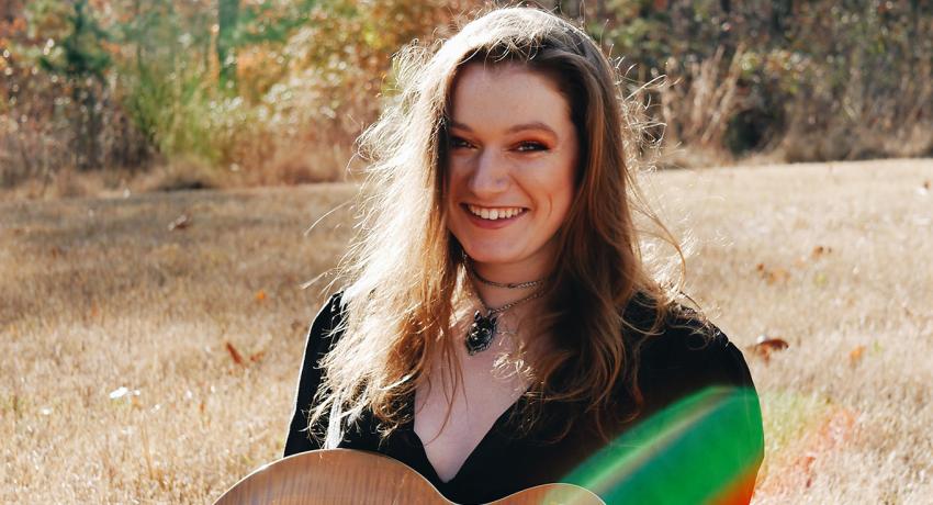 Musician Brianna Bollinger holding one of her guitars, sitting in the grass, with trees behind her